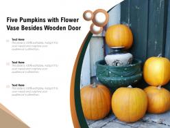 Five pumpkins with flower vase besides wooden door